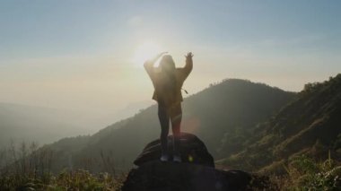 Sarı ceketli bir kadının güneşin doğuşunda dağın zirvesinde durup kollarını kaldırarak temiz hava soluyarak sakin bir günün tadını çıkardığını hayal edin. bio-turizm