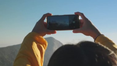 Sırt çantasıyla gezen bir kadın güzel dağ anılarını ve deneyimlerini kaydetmek için akıllı telefon kullanıyor. Mutlu ve heyecanlı genç kadın açık hava macerasında