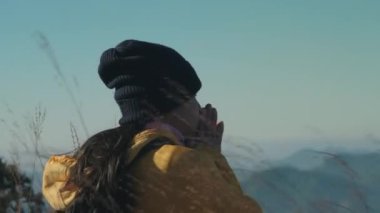Dağın tepesinde sıcak kalmaya çalışan Asyalı bir kadın turistin portresi. Sarı ceketli ve sırt çantalı örgü şapkalı turist kadın ellerini ısıtıyor. Üşüyorum.