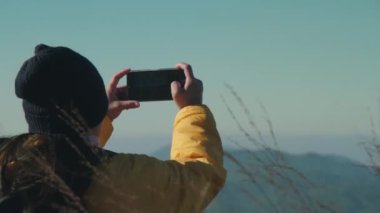 Sırt çantasıyla gezen bir kadın güzel dağ anılarını ve deneyimlerini kaydetmek için akıllı telefon kullanıyor. Mutlu ve heyecanlı genç kadın açık hava macerasında