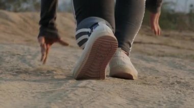 Akşam gün batımında köy yolunda koşan genç bir kadın. Toprak bir yolda koşan atletik ayakkabılar giyen bir sporcunun ayaklarına yakın plan..