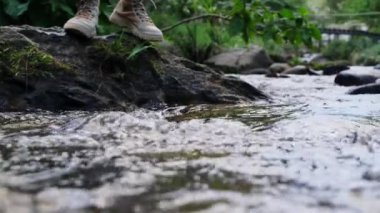 Dişi gezgin, ormandaki bir derenin karşısındaki bir kayanın üzerinde yürüyor. Kahverengi ayakkabılı bacakları sığ suda yürürken görmek. Macera, keşif ve seyahat kavramı.