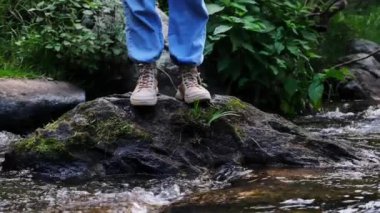 Dişi gezgin, ormandaki bir derenin karşısındaki bir kayanın üzerinde yürüyor. Kahverengi ayakkabılı bacakları sığ suda yürürken görmek. Macera, keşif ve seyahat kavramı.
