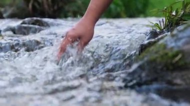 Kadın eli nehirde soğuk suya dokunuyor. Seyahat eden kadın bir kayanın üzerinde duruyor ve elleriyle bir dereye dokunuyor. Elin yakın çekimi.