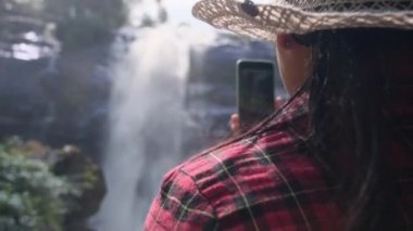 Hasır şapkalı ve kırmızı ekose gömlekli mutlu hippi kadın akıllı telefon kullanarak Chiang Mai kuzey Tayland 'daki Doi Inthanon Ulusal Parkı' ndaki Wachirathan şelalesinde doğanın tadını çıkarıyor. Tayland Seyahat Konsepti