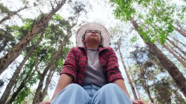 Hasır şapkalı ve kırmızı ekose gömlekli mutlu bir hippi kadının düşük açılı görüntüsü çam ağaçlarının altında oturup doğanın tadını çıkarıyor. Mutlu Asyalı kadın gezgin çam ağacı bahçesini geziyor..