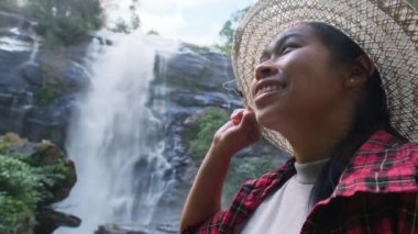Hasır şapkalı ve kırmızı ekose gömlekli mutlu hippi kadın Chiang Mai kuzey Tayland 'daki Doi Inthanon Ulusal Parkı' ndaki Wachirathan şelalesinde doğanın tadını çıkarıyor. Tayland Seyahat Konsepti