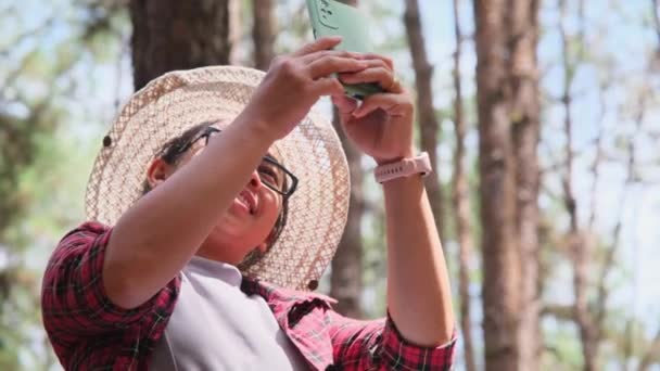 松の木の下に座って テントの背景に秋の森の中でスマートフォンを使用して美しいアジアの女性 自然の中でキャンプ — ストック動画