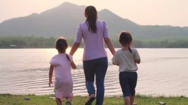 Mutlu anne ve iki tatlı kız gün batımında göl kenarında el ele yürüyorlar. Aile tatillerini birlikte geçiriyor..