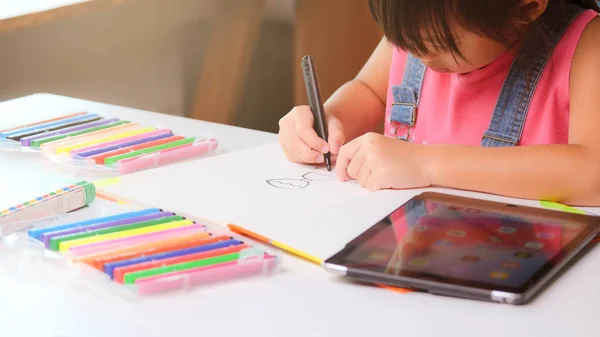 Elinde sihirli kalemle dijital tablet ve kırtasiye malzemelerini kağıda çizen sevimli küçük bir kız. Çocuk internette ev ödevi için dijital tablet arama bilgileri kullanıyor, evde eğitim.