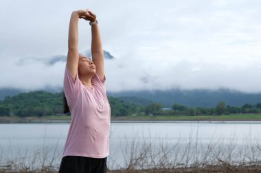 Asian woman warming up and stretching her arms in the riverside countryside in the morning before jogging. Sports and recreation healthy lifestyle concept.