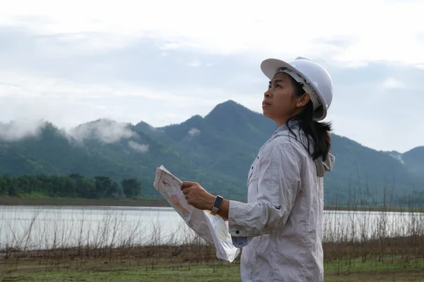 Engineering ecologist woman in a helmet holding a blueprint stands on the bank of a river to develop a hydroelectric dam to generate electricity. Clean energy and Technology concepts.