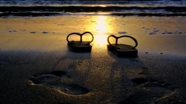 Sandals on the beach on a beautiful background of the beach at sunset with golden light reflecting on the water surface and soft waves. Summer vacation and travel concept.