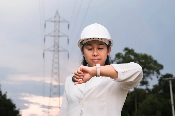 Asian female electrical engineer looking at a smart watch on the background of sky and high voltage poles.