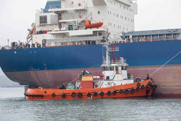 Red tug boat assisting huge ship in port — Stock Photo, Image