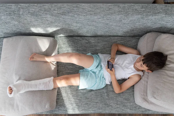 小男孩腿断了石膏 躺在家里的沙发上 用手机玩 — 图库照片