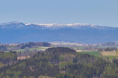 Manzara. Yeşil orman. Tepeler. Tarlalar. Karla kaplı dağ zirveleri. Küçük evler. Mavi gökyüzü