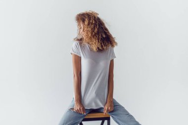 Beyaz, boş tişörtlü, gür kıvırcık saçlı, gülümseyen kadın beyaz arka planda oturuyor. Model.