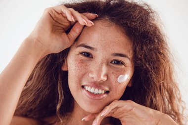 Nemlendirici yüz kremi kullanan gür kıvırcık saçlı ve çilli bir kadının portresi..