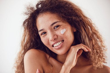 Nemlendirici yüz kremi kullanan gür kıvırcık saçlı ve çilli bir kadının portresi..