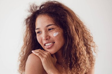 Nemlendirici yüz kremi kullanan gür kıvırcık saçlı ve çilli bir kadının portresi..