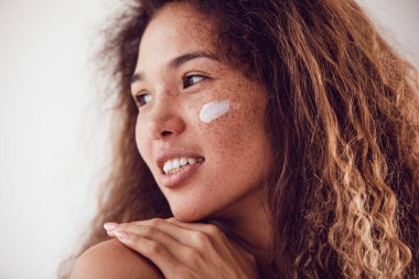 Nemlendirici yüz kremi kullanan gür kıvırcık saçlı ve çilli bir kadının portresi..
