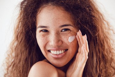 Yüzüne nemlendirici süren kıvırcık saçlı ve çilli kadın portresi.