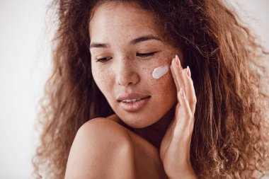 Yüzüne nemlendirici süren kıvırcık saçlı ve çilli kadın portresi.