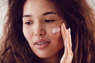 Yüzüne nemlendirici süren kıvırcık saçlı ve çilli kadın portresi.