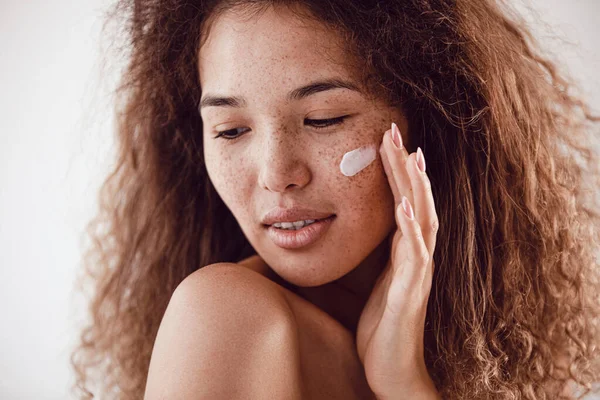 Yüzüne nemlendirici süren kıvırcık saçlı ve çilli kadın portresi.