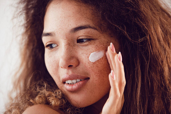 Portrait of woman with curly hair and freckles applying moisturizer to her face.