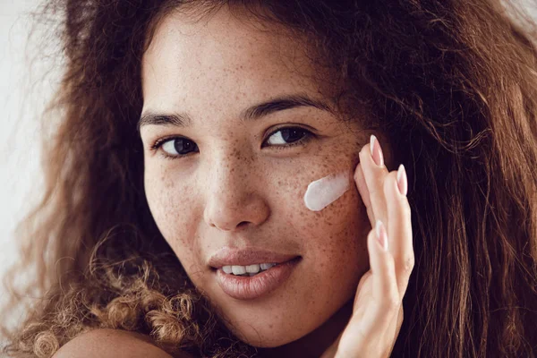 Yüzüne nemlendirici süren kıvırcık saçlı ve çilli kadın portresi.