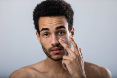 Genç Afrikalı Amerikalı adam yüzüne nemlendirici sürmüş. Erkek cilt bakımı.