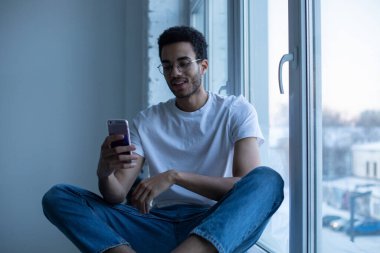 Gözlüklü ve beyaz tişörtlü Afro-Amerikalı adam pencerenin yanında otururken akıllı telefon kullanıyor..