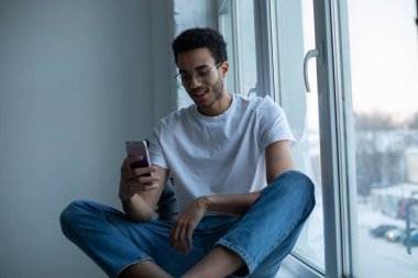 Gözlüklü ve beyaz tişörtlü Afro-Amerikalı adam pencerenin yanında otururken akıllı telefon kullanıyor..