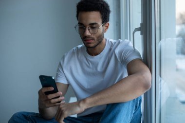 Gözlüklü ve beyaz tişörtlü Afro-Amerikalı adam pencerenin yanında otururken akıllı telefon kullanıyor..