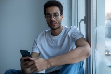 Gözlüklü ve beyaz tişörtlü Afro-Amerikalı adam pencerenin yanında otururken akıllı telefon kullanıyor..