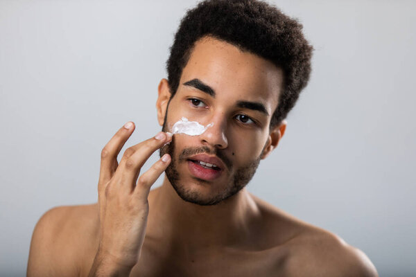 Young african american man applies moisturizer on his face. Male skin care.