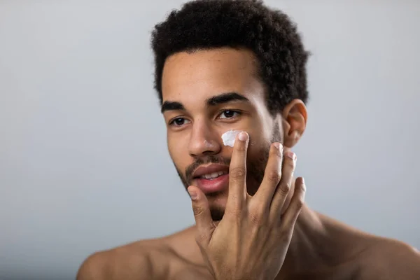 Genç Afrikalı Amerikalı adam yüzüne nemlendirici sürmüş. Erkek cilt bakımı.