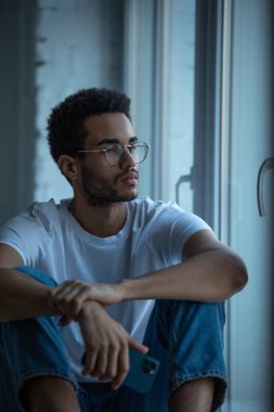 Çekici Afro-Amerikan gözlüklü ve beyaz tişörtlü adam pencere eşiğinde oturuyor..