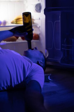 Bir adam açık buzdolabının yanında yatar ve şişeden şarap içer..