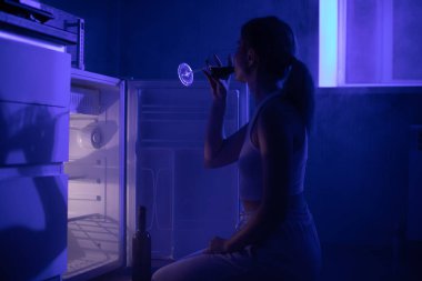 Üzgün kadın şarap içiyor açık bir buzdolabının önünde karanlıkta oturuyor..
