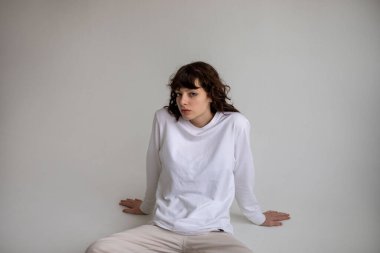 Beyaz uzun kollu tişörtlü kıvırcık saçlı genç bir kadın beyaz arka planda oturuyor. Model.