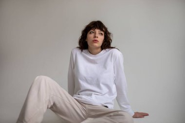 Beyaz uzun kollu tişörtlü kıvırcık saçlı genç bir kadın beyaz arka planda oturuyor. Model.