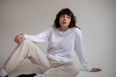 Beyaz uzun kollu tişörtlü kıvırcık saçlı genç bir kadın beyaz arka planda oturuyor. Model.