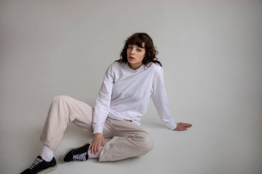 Beyaz uzun kollu tişörtlü kıvırcık saçlı genç bir kadın beyaz arka planda oturuyor. Model.