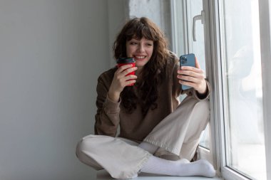 Elinde bir fincan kahveyle genç ve çekici bir kadın pencere kenarında oturmuş selfie çekiyor..