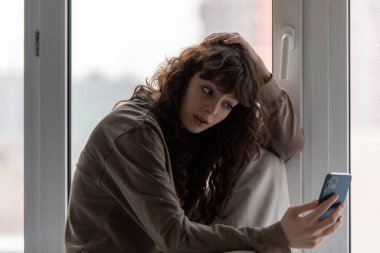 Genç çekici kadın pencerenin kenarında otururken akıllı telefon kullanıyor..