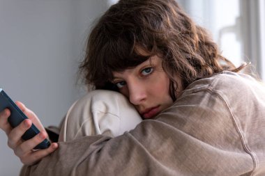 Pencerenin yanında otururken akıllı telefon kullanan kıvırcık bir kızın portresi.