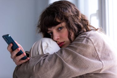 Pencerenin yanında otururken akıllı telefon kullanan kıvırcık bir kızın portresi.